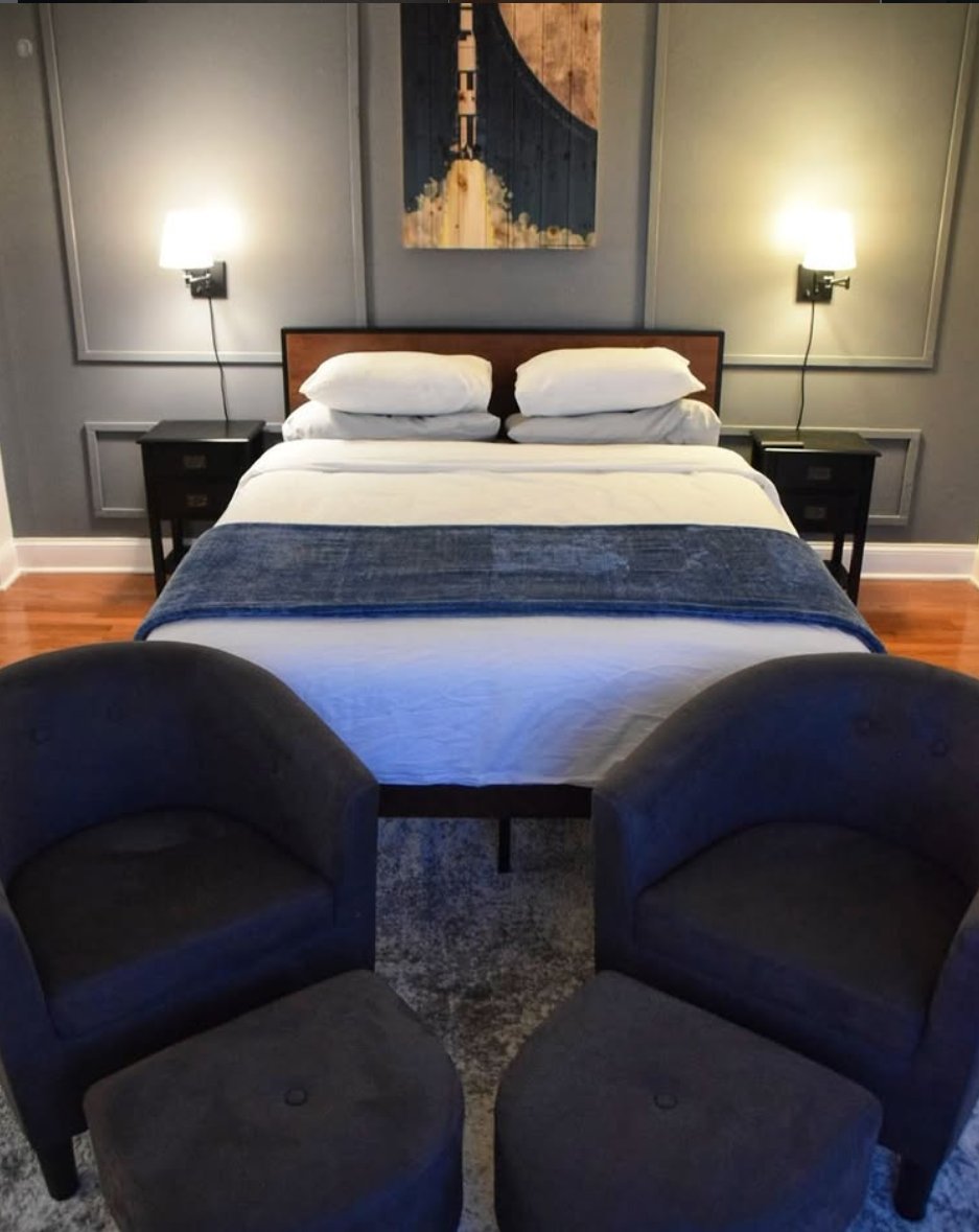 Bedroom with grey paneled wall, neatly made bed, and armchairs